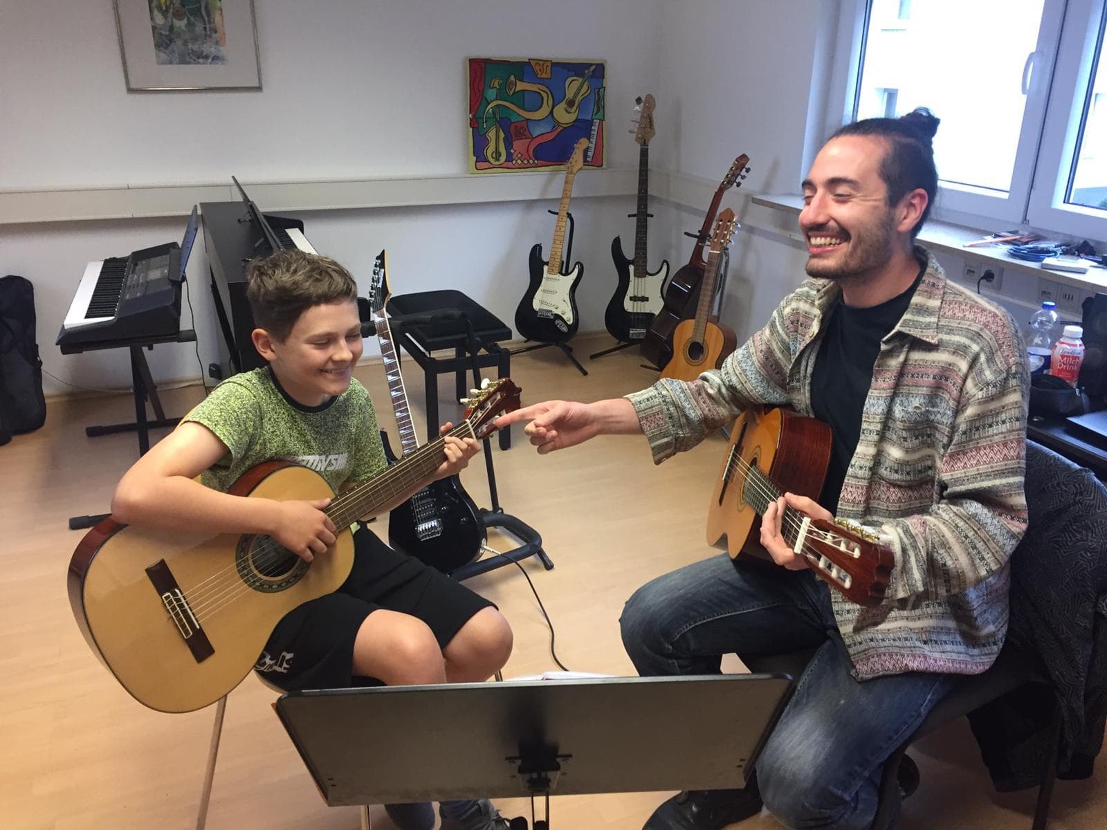 Ein Lehrer zeigt einem Jungen einen Griff auf der Gitarre, beide lachen.