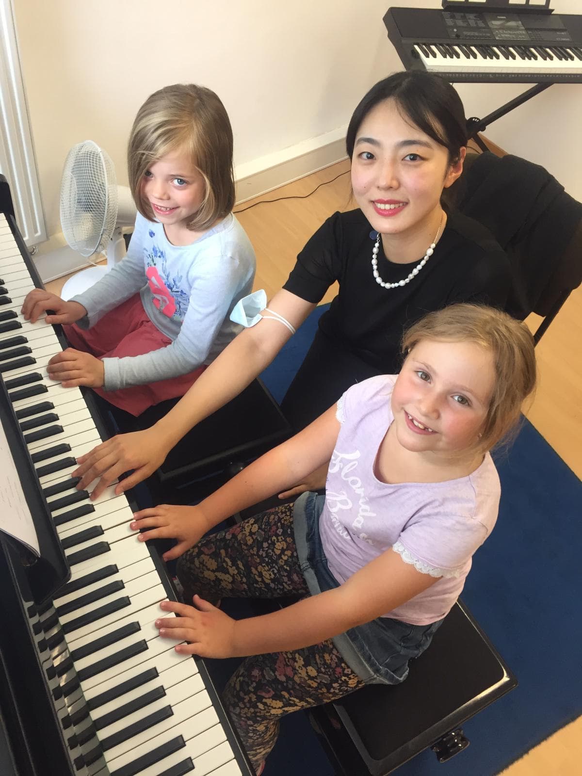 Zwei Mädchen sitzen mit ihrer Lehrerin am Klavier und lächeln.